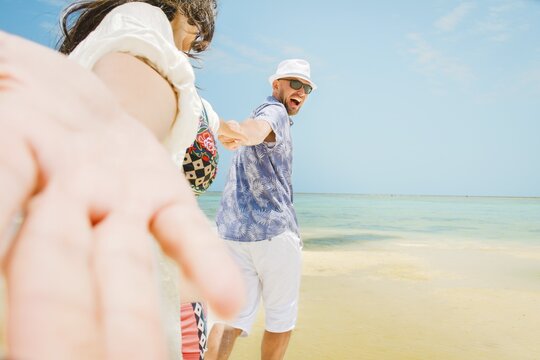 Cheerful young man leading woman couple holding hands run to beach have fun together on shallow turquoise ocean water sandy beach tropical island resort destination. Relationship honeymoon vacation