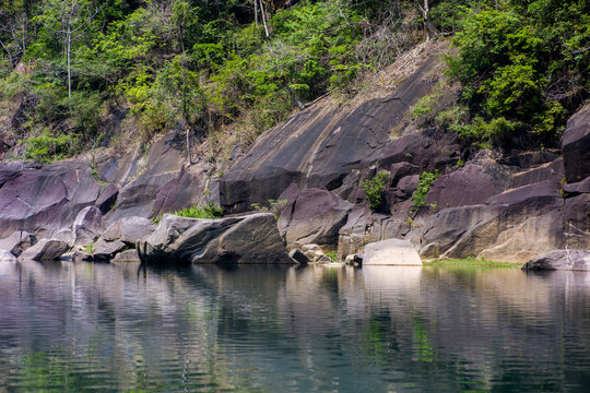 Meghalaya, India - 31 March 2023: View of the rocky riverbank with dark stone formations and lush green forest foliage reflecting in the calm water.