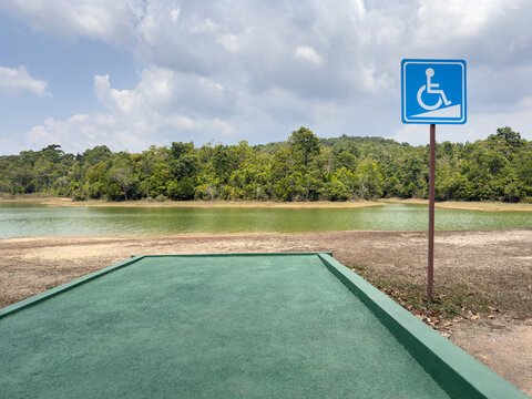 green, textured concrete viewing platform leads toward a calm, murky green lake, bright blue wheelchair accessibility sign mounted on pole, pictogram of a person in a wheelchair navigating a ramp