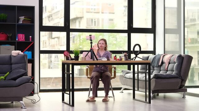 Cheerful teenage schoolgirl uses modern smartphone to record video blog. Creative little blogger sits at desk and creates entertaining vlog at home