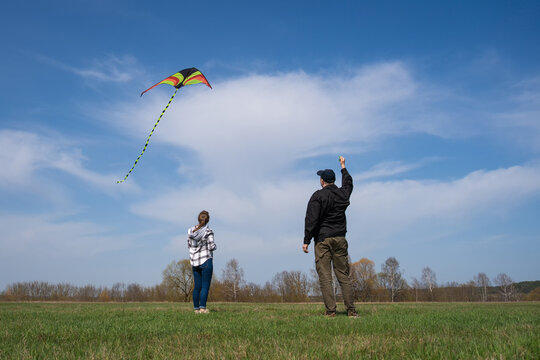 People standing in a green field, a man holding a kite string with a colorful kite flying high in the blue sky, while a woman looks up and observes the outdoor recreation