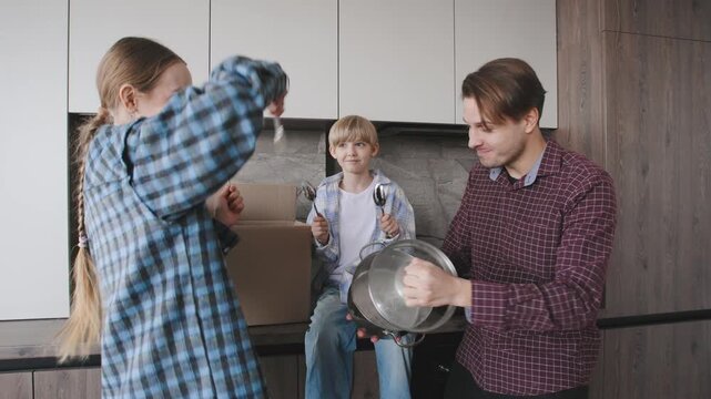 A family stands in the kitchen surrounded by cardboard boxes, playing with kitchenware and utensils. They are having fun unpacking their belongings in their new apartment.