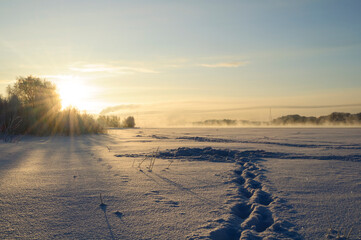 Winter landscape sunrise background. Bright sun rising over a snow covered field. Footprints and tire tracks across the snow with mist drifting over the distant horizon © syntheticmessiah