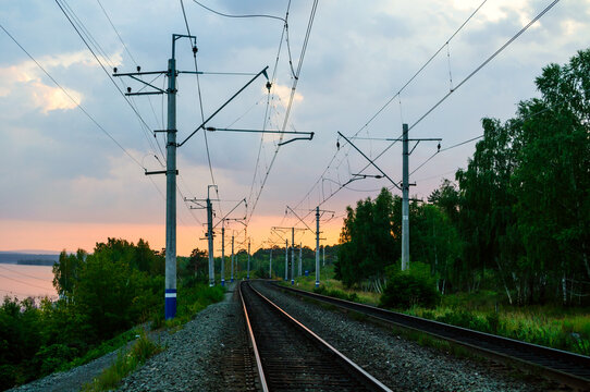 Railway landscape background. Curving train tracks leading into the distance at sunset. Overhead power lines, concrete poles and green trees along the route