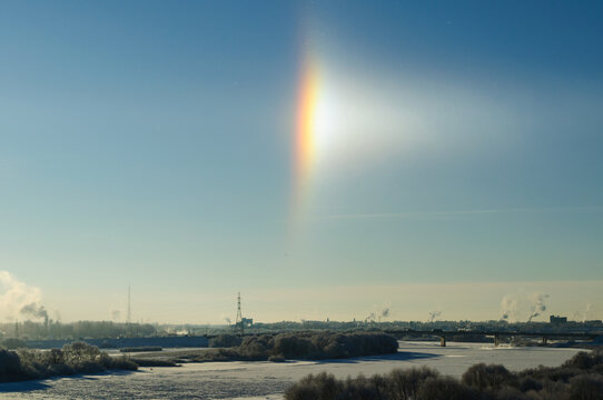 Winter industrial landscape background. A bright sun halo in a clear blue sky above a frozen river. Frosted trees, a bridge and smoking chimneys on the horizon
