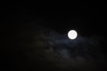 Fototapeta premium Midnight sky background. A bright white full moon glowing in a dark night sky. Thin clouds drifting across the sky around the bright lunar disc