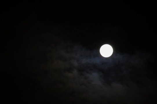Midnight sky background. A bright white full moon glowing in a dark night sky. Thin clouds drifting across the sky around the bright lunar disc