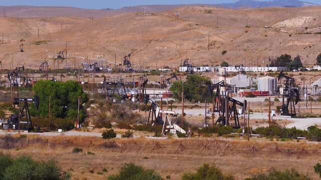 Aerial view pans across active oil pump jacks and industrial drilling infrastructure operating on an arid California oilfield surrounded by barren hills. Oil tariffs, trade war, price plunge.