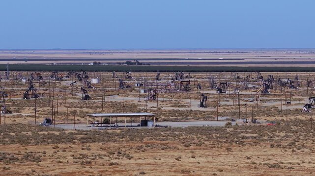 Aerial view of an expansive California oil field filled with operating pump jacks, wells, pipelines, and utility poles across an arid landscape. Oil tariffs, trade war, price plunge.