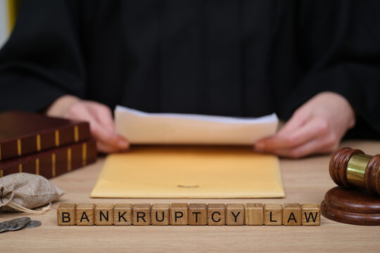 A judge dressed in black robes sits at a wooden desk, hands resting on a yellow folder.