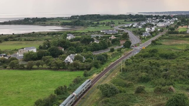 A high angle drone shot captures a modern passenger train traveling along tracks parallel to a coastal road in a lush Irish village, showcasing sustainable public transport.
