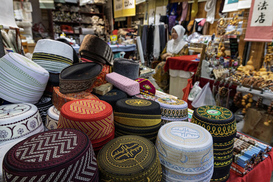 Xi'an, Shaanxi, China - 14 September 2026: View of traditional embroidered Muslim caps at a market stall, with a woman sitting in the background surrounded by wooden crafts.
