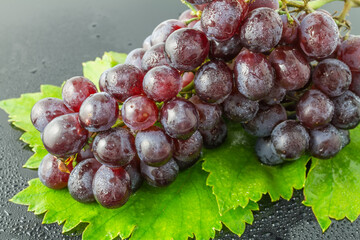 A close-up of a fresh red grape cluster with glistening water droplets, resting on green grape leaves against a dark surface. © tai