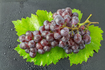 A cluster of fresh, glossy purple grapes rests on vibrant green vine leaves, with water droplets adding a crisp, fresh appearance. © tai