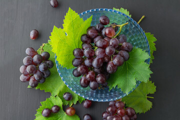 Fresh ripe red grapes arranged in a blue glass bowl with vine leaves, placed on a dark surface. © tai