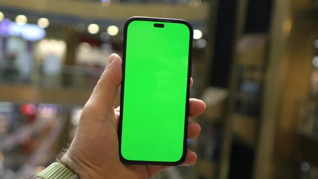 Hand holds a smartphone with a green screen. The mall background is softly blurred. The phone display shows a blank screen. This scene is calm and professional. Ideal for marketing demos