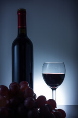 Fototapeta premium A still life composition featuring a red wine bottle, filled wine glass, and fresh grape cluster against a dark studio background.