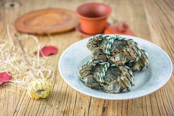 A still life of raw tied crabs resting on a light plate, placed on a rustic wooden table with ceramic tableware and decorative elements. © tai