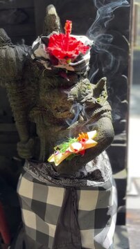 A stone statue of a deity holds offerings with smoke rising. The statue is adorned with a red flower and wears a traditional black and white checkered cloth.
