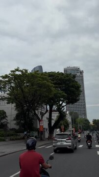 Urban street scene with tall buildings and green trees. A car drives along the road under a cloudy sky. City life and architecture are evident.