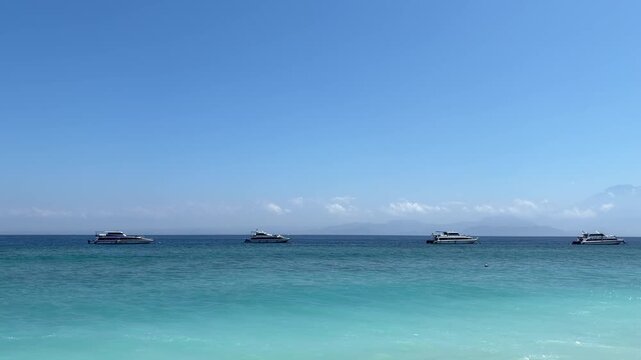 A clear blue sky over a tranquil sea with several boats in the distance. The water is a gradient of turquoise and deep blue, creating a serene coastal scene.