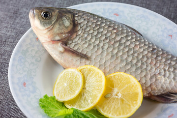 A whole raw crucian carp, presented with lemon slices and a lettuce leaf on a decorative plate, prepared for cooking. © tai