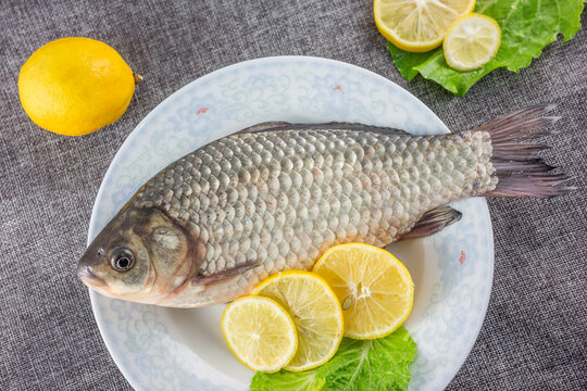 A whole raw crucian carp arranged with lemon slices and lettuce on a light plate, prepared for cooking or serving.
