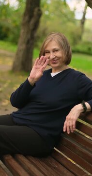 Vertical video of beautiful smiling mature blond woman sitting on the bench in park