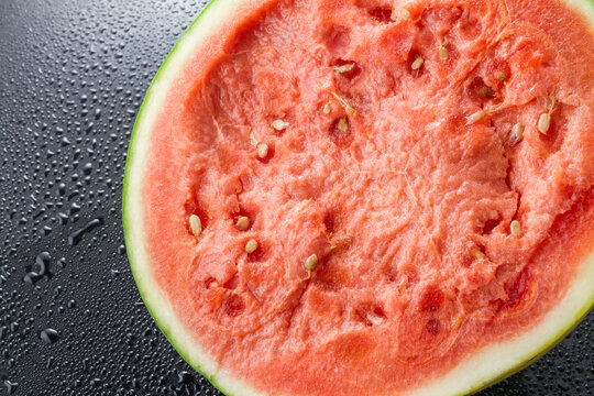 A close-up of a sliced ripe watermelon with seeds, resting on a wet dark surface with water droplets.