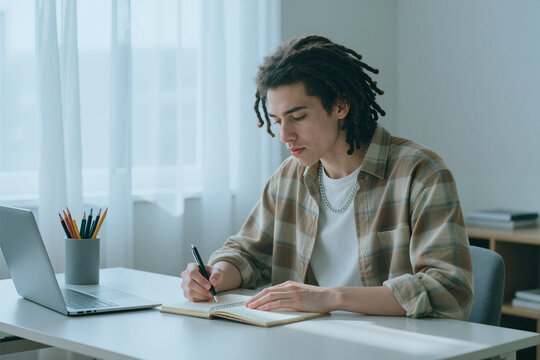Caucasian young adult man with dreadlocks writing in notebook at desk with laptop, studying or working from home. Image suitable for remote education and hybrid work campaigns