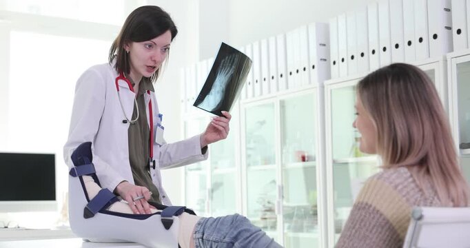 Female doctor reviews leg X-ray image with woman during clinic consultation. Physician holds radiograph explaining medical findings during appointment