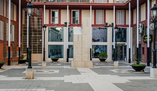 Malaga, Andalusia, Spain - 18 May 2020: View of a quiet modern courtyard featuring a central stone monument, paved ground, and surrounding red and cream buildings with street lamps.