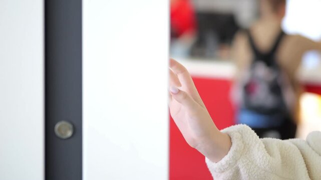 Close-up of a person opening a white door. Background shows blurred indoor space with people. Hand pulls door open slowly. Focus stays on hand and door handle. Ambient lighting with warm tones