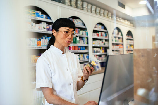 Pharmacist checking medicine bottle at computer in pharmacy
