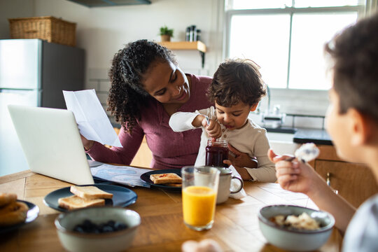 Mom works on laptop while helping toddler with jam at breakfast