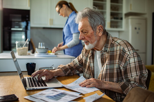 Senior man paying bills on laptop while caregiver prepares meal