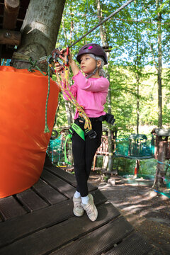 Young girl in a safety helmet and harness navigates a rope adventure park in a sunny forest. The scene illustrates active childhood, physical development, bravery, and healthy outdoor recreation.