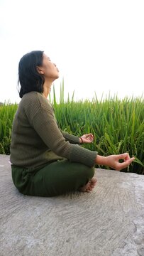 A woman meditating in a peaceful lotus position outdoors near a lush green rice field.