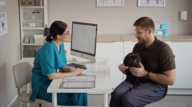 Caucasian female veterinarian consulting young adult Caucasian male holding black cat in clinic office. Clip useful for pet healthcare, veterinary advertising, animal wellness campaigns