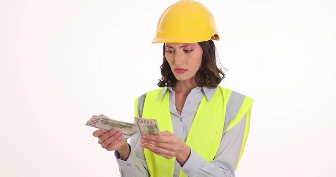 Frustrated female construction worker counts stack of dollar bills. Woman disappointed by unexpectedly low paycheck against white background