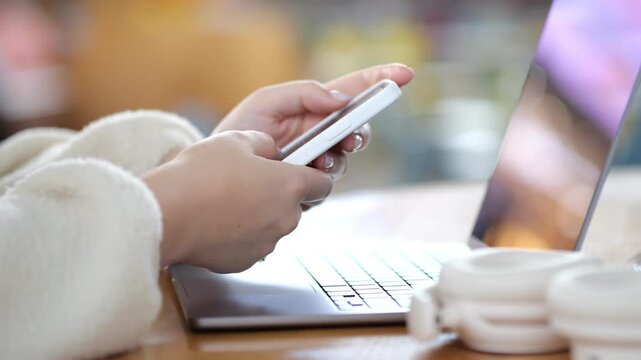 Close-up hands holding smartphone. Laptop rests nearby on wooden table. Warm, cozy indoor setting with blurred background. Person wears soft white sweater. Two coffee cups sit beside laptop