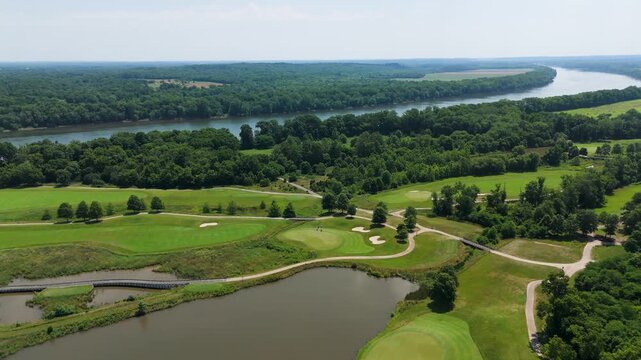 Aerial drone tracking shot of a beautiful lush green golf course beside a wide river on a clear summer day. Features manicured fairways, sand traps, cart paths, and bridges over wetlands.
