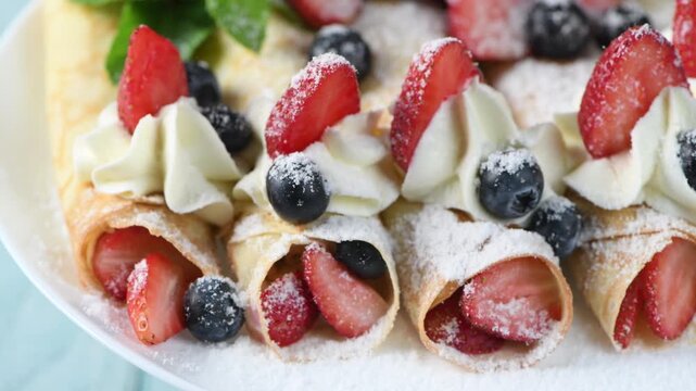 Thin pancakes filled with fresh berries, topped with whipped cream, strawberries, and blueberries, dusted with powdered sugar and mint. Close-up of the dessert on a white plate. Horizontal video. Circ