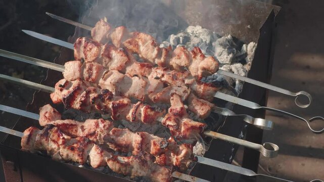 Juicy pieces of meat are grilling on metal skewers over hot charcoals in a brazier. Fragrant smoke rises from the roasted shish kebab during outdoor cooking
