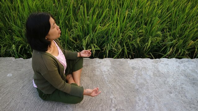 A young Asian woman practicing meditation and mindfulness in a serene rice field setting.