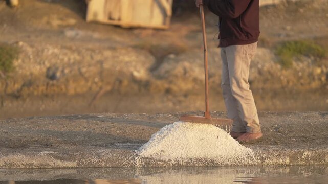natural sea salt harvesting in indonesia, farmer using wooden rake tool in salt field, pond, pan, traditional farming, southeast asia, slow motion
