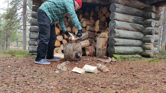 A man wearing plaid shirt and black pants is splitting logs with an axe next to a wooden shed in the Lapland forest