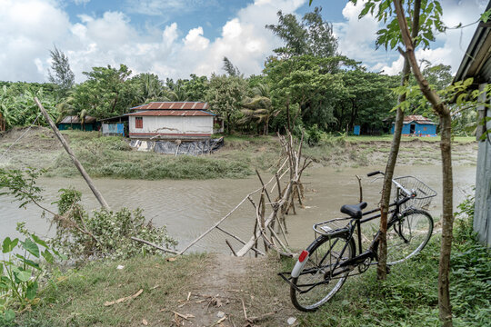 Noakhali, Chittagong Division, Bangladesh - 15 August 2026: View of a makeshift wooden bridge crossing a river with a bicycle in the foreground and rural houses among trees under a cloudy sky.