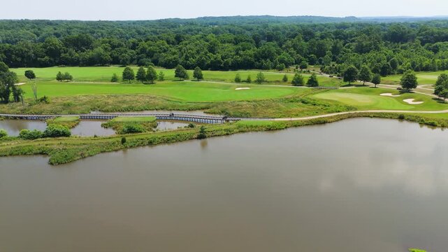 Aerial drone tracking shot moving right over a golf course water hazard on a sunny summer day. Features a long wooden bridge crossing a pond, lush green fairways, sand traps, and a dense forest.