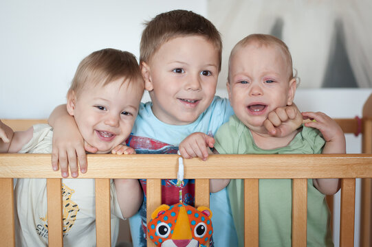 Three cheerful children, three brothers, are standing closely together in a nursery, smiling and playing with toys. The bright space reflects their playful energy and bond as siblings.